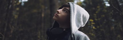 teen boy with a hoodie and headphones looks up at the trees. 