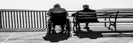 Black and white. Two elderly people sit on a bench and in a wheelchair overlooking the ocean