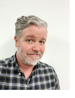 Headshot of Brandon: A man with wavy silver hair and a plaid shirt smiles at the camera